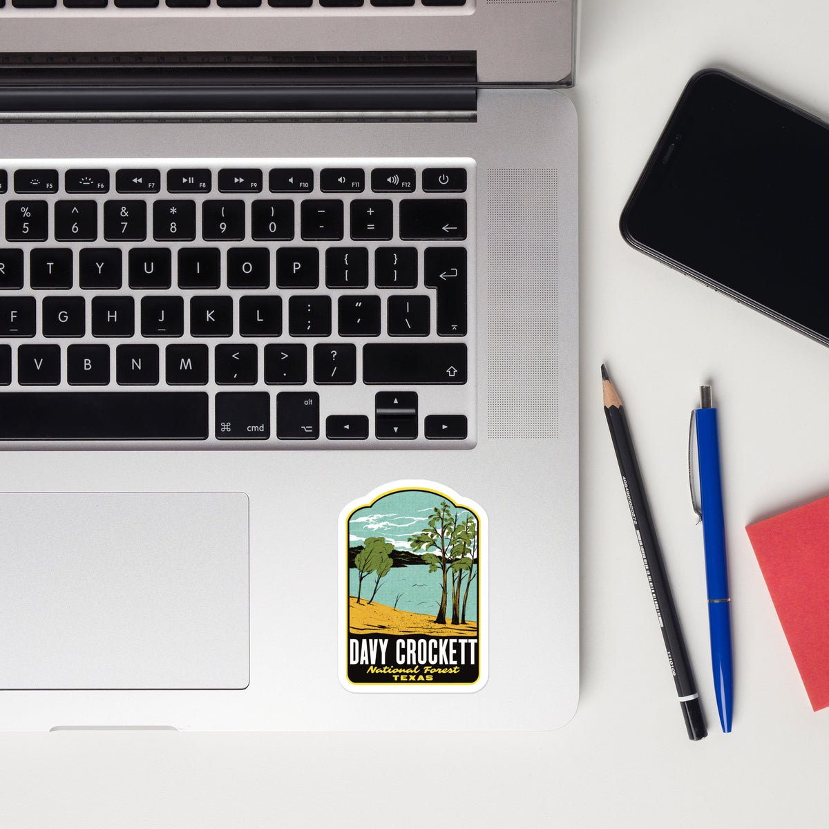 A rectangular sticker featuring a retro landscape illustration of Day Crockett, Texas, placed on the palm rest of an open silver laptop. The sticker depicts a lake, trees, and a yellow beach.