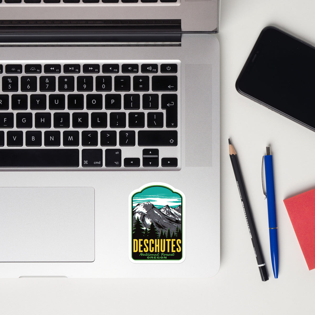Laptop with a ' Deschutes National Forest' sticker, phone, pencil, and pen on a white surface