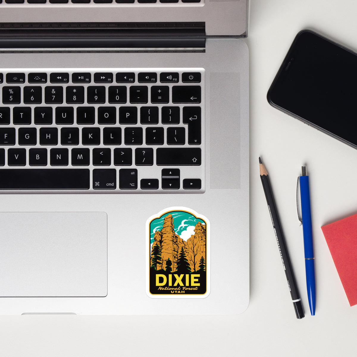 A rectangular sticker with rounded corners featuring a scenic mountain and pine tree illustration above the word 'DIXIE' in yellow, placed on the lower right corner of a silver laptop keyboard. The laptop sits on a white desk alongside a black smartphone, a pencil, a blue pen, and a red notebook.