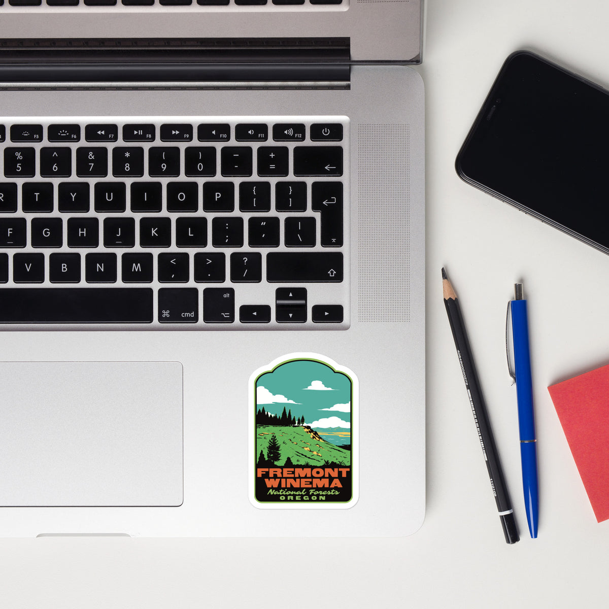 A rectangular die-cut sticker featuring a retro-style scenic illustration of Fremont Winery in Napa Valley, applied to the palm rest of a silver laptop. The scene is styled as a flat-lay on a white desk with a smartphone, a pencil, a blue pen, and a red notebook.