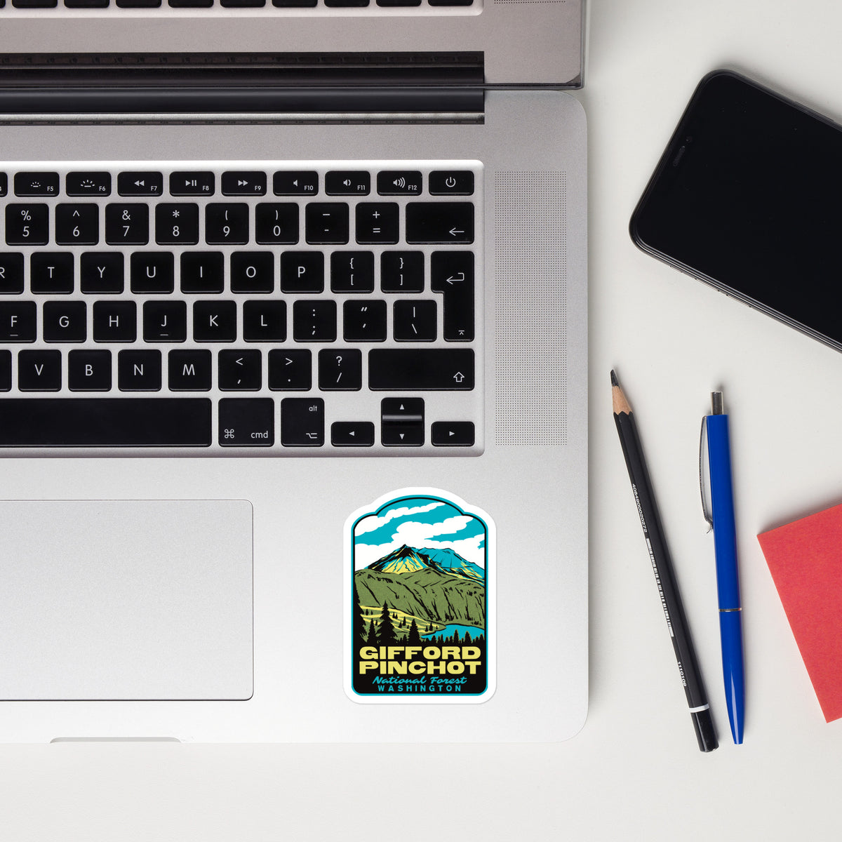 A rectangular die-cut sticker with a mountain landscape illustration and the text 'GIFFORD PINCHOT National Forest' is placed on the palm rest of a silver laptop. The scene is styled as a flat lay on a white desk with a black smartphone, a pencil, a blue pen, and a red notebook.