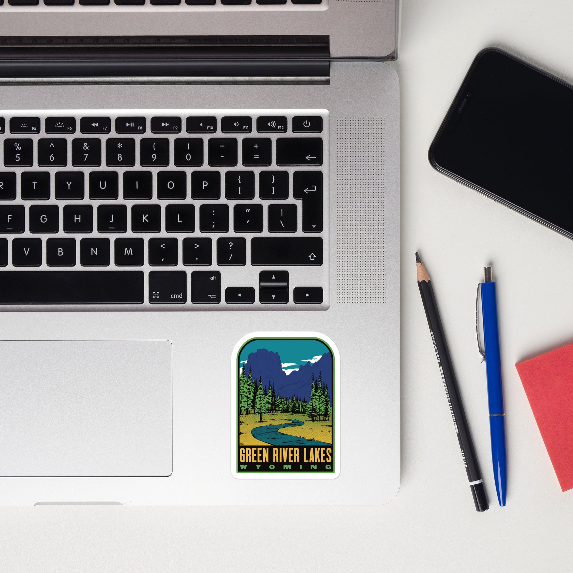 Laptop with a 'Green River Lakes, Wyoming' sticker, phone, pencil, pen, and notebook on a white surface