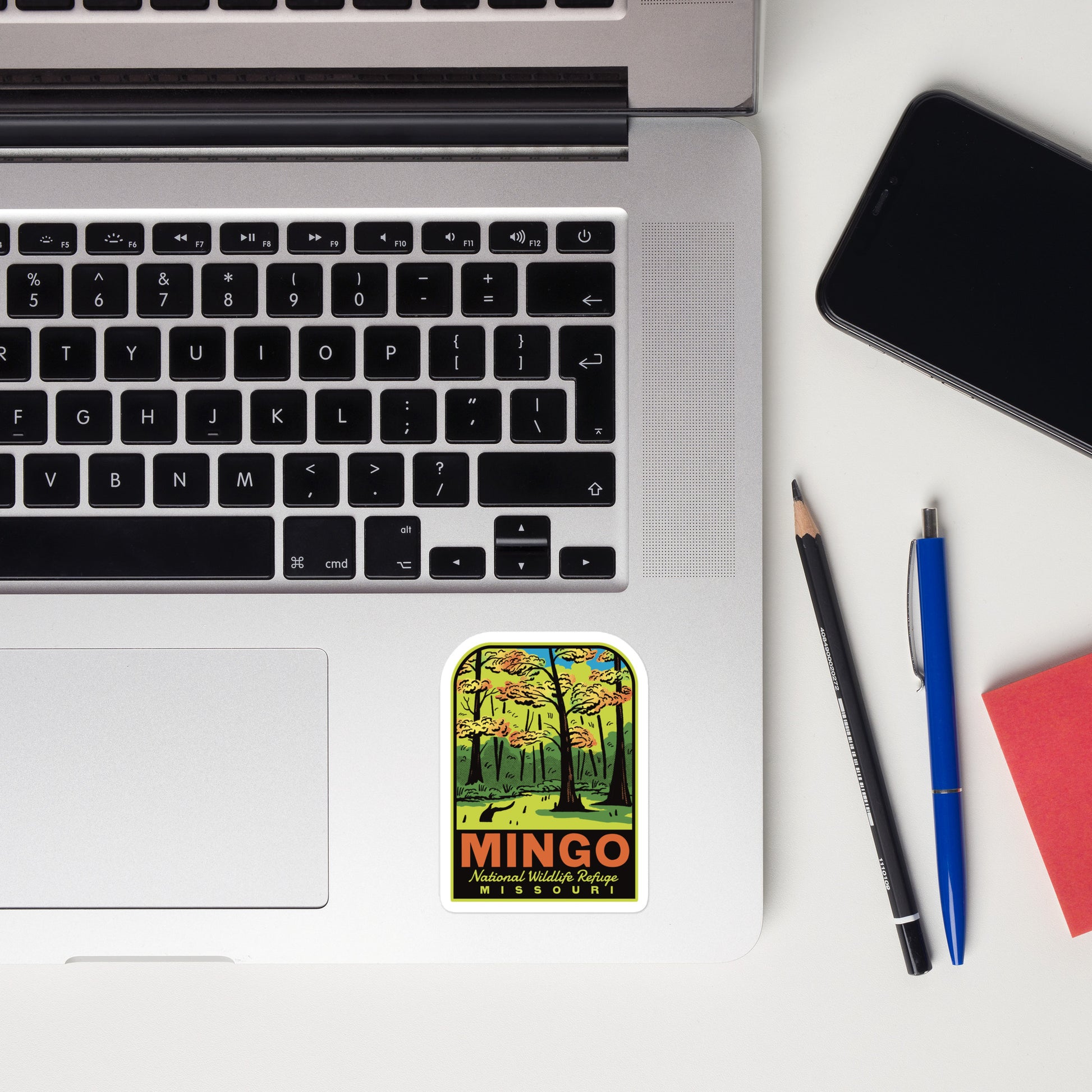 Laptop with a 'Mingo National Wildlife Refuge' sticker, phone, pencil, pen, and notebook on a white surface.