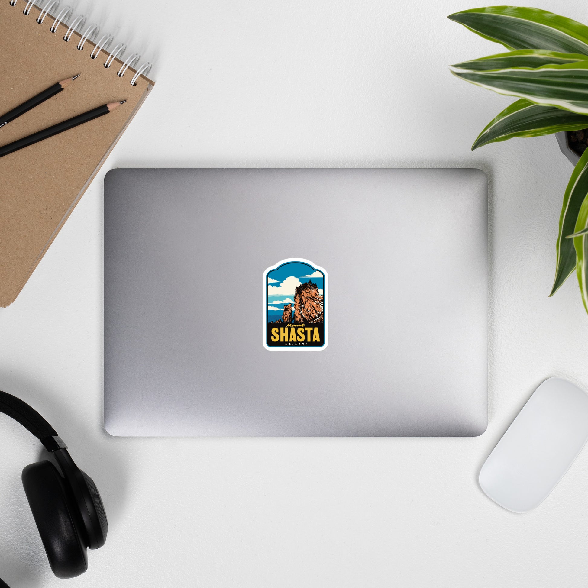 Laptop with a colorful Mount Shasta sticker on a white surface with a notebook, pencils, and plant.