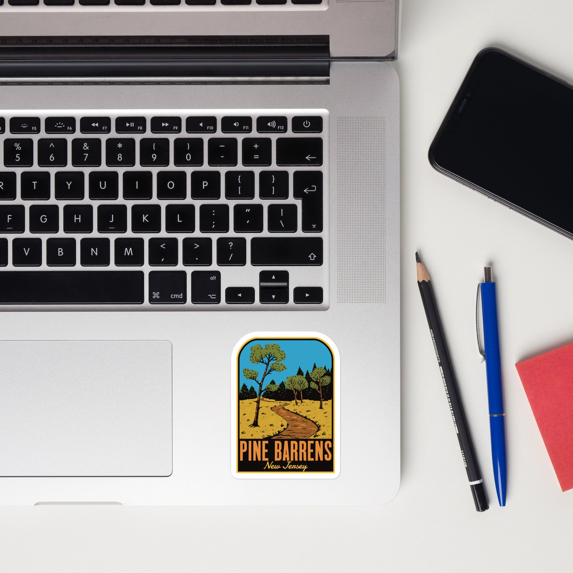 Laptop with a 'Pine Barrens, New Jersey' sticker, phone, pencil, pen, and notebook on a white surface.