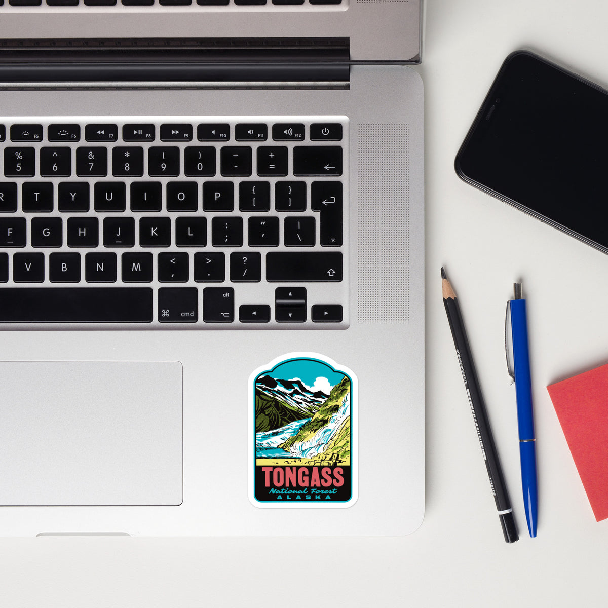 A rectangular sticker with rounded corners featuring a colorful mountain landscape and the text 'TONGASS' and 'Alaska', placed on the lower left corner of a silver laptop. The laptop sits on a white desk alongside a black smartphone, a pencil, a blue pen, and a red notebook.